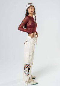 Top de malla de manga larga color marrón con bordado floral, combinado con pantalones cargo color crema que presentan una gráfica de geisha, y zapatillas beige con detalles en verde.