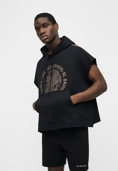 Tânăr purtând un hanorac fără mâneci negru cu un grafic bej și pantaloni scurți negri cu logo-ul alb Les Benjamins.