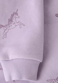 Tessuto colore lilla chiaro con motivo unicorno in tonalità viola più scure. Il polsino è a coste, aggiungendo texture e profondità al capo.