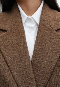 Femme portant une chemise blanche avec col sous un blazer marron à motif chevrons avec larges revers.