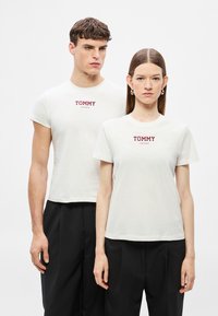 Άσπρα βαμβακερά μπλουζάκια με στρογγυλές λαιμόκοψη, με κόκκινο λογότυπο "TOMMY JEANS" στο στήθος. Το μοντέλο φοράει μαύρο παντελόνι.