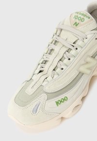 Beżowe i jasnoróżowe sneakersy New Balance 1000 z kremowymi sznurowadłami, haftowanym zielonym logo oraz detalami z siateczki i zamszu na białym tle.