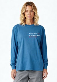 Chemise en coton bleu à manches longues avec texte en blanc et rouge. Coupe décontractée avec encolure ronde et poignets côtelés. Texture douce.