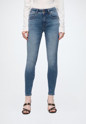 Donna che indossa jeans skinny azzurri chiari, top bianco a maniche lunghe e sandali neri con tacco alto, in piedi davanti a uno sfondo semplice.