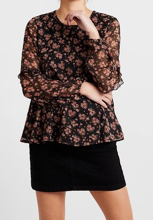 Mujer con una blusa negra transparente de manga larga con un patrón floral rosa y una falda negra, con los brazos cruzados y una mano cerca del mentón.