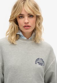 Sudadera gris con bordado de "Superdry Athletics Beach Club" en azul marino. Cuenta con cuello redondo, corte relajado y textura suave.