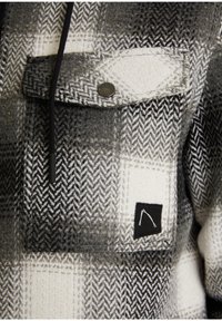 Gros plan sur la poche d'une veste en tissu à chevrons gris et blanc, avec un bouton-pression métallique et une petite étiquette noire ornée d'un motif blanc.