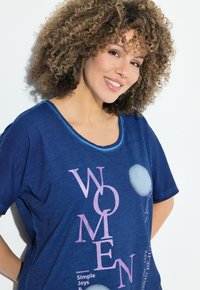 Usměvavá žena s kudrnatými vlasy, oblečená v modré košili s růžovým nápisem "WOMEN" a dekorativními bílými kruhy, stojící proti bílému pozadí.