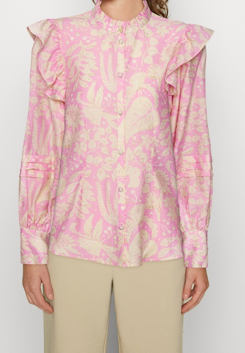 Blouse rose à motif floral avec épaules à volants, fermeture à boutons et manches longues. Le tissu présente une texture lisse avec un léger éclat.