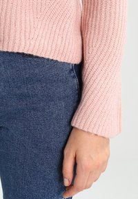 Main reposant le long du corps, vêtue d'un pull en maille rose et d'un jean en denim bleu, sur un fond clair uni.