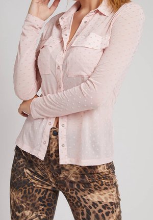 Osoba ubrana w przezroczystą jasnoróżową bluzkę z długim rękawem z teksturowanymi kropkami oraz spodnie w panterkę, pozująca z założonymi skrzyżowanymi rękami.