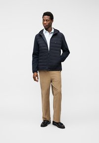 Άνδρας που στέκεται, φοράει σκούρο μπλε μπουφάν με επένδυση και κουκούλα, λευκό πουκάμισο, μπεζ παντελόνι και μαύρα παπούτσια, κοιτάζει προς τα πλάγια με το ένα χέρι στην τσέπη.