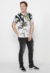 Camiseta blanca de manga corta con salpicaduras en estilo graffiti en negro y verde, combinada con pantalones cargo negros y zapatillas negras con acentos blancos.