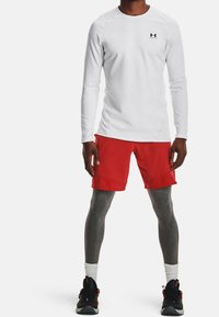 Weißes langärmliges Sportshirt mit Logo, kombiniert mit roten Shorts und grauen Leggings, ergänzt durch schwarze und rote Turnschuhe.