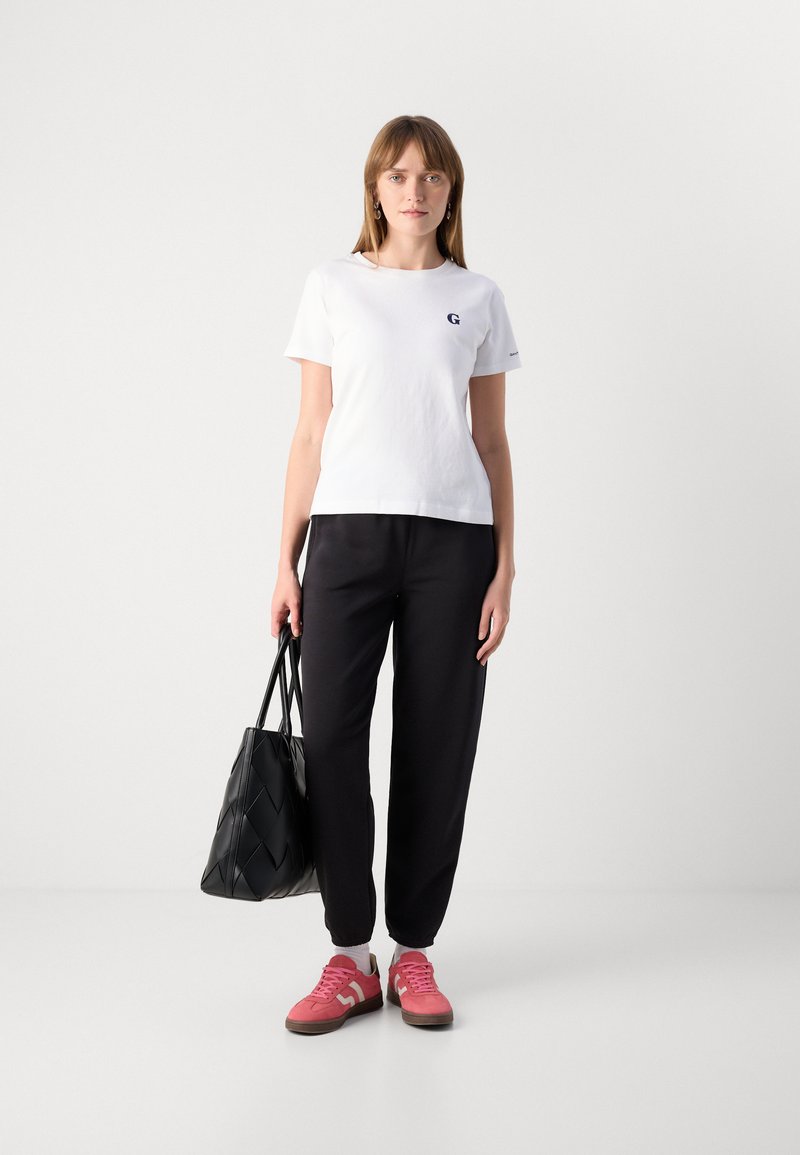 Weißes Baumwoll-T-Shirt mit einem kleinen blauen Logo, kombiniert mit schwarzen Jogginghosen und pinken Sneakers. Hält eine schwarze gewebte Tragetasche.