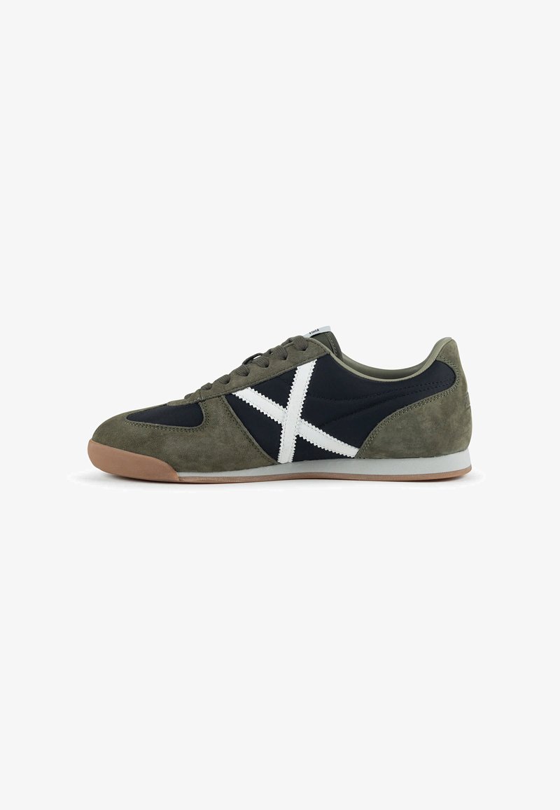 Sneaker profil bas en daim vert olive, panneaux en tissu noir, design X cousu blanc, semelle en caoutchouc marron et fermeture à lacets sur fond uni.