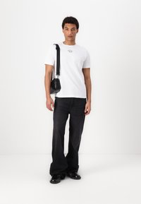 Hvid bomulds t-shirt med logo, sorte løsning bukser og sort skuldertaske. Modellen bærer sorte sko. Ensfarvet hvid baggrund.