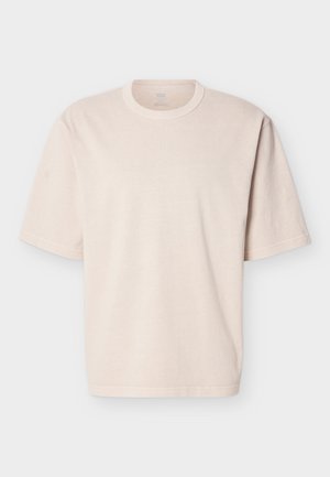 Beige oversized T-skjorte med korte ermer, rund hals og enkel søm, vist flatt på hvit bakgrunn.