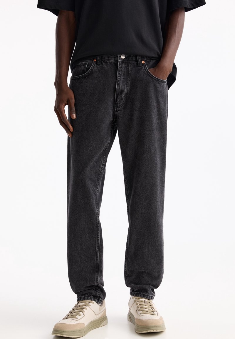 Homme portant un jean noir ample avec des poignets élastiques et des baskets beiges, debout devant un fond blanc uni.