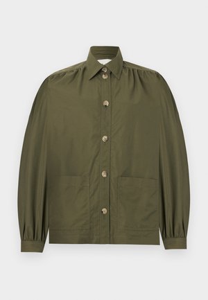 Moteris dėvi alyvuogių žalios spalvos užsegamą švarką ant kreminės spalvos aukštu kaklu megztinio ir spalvotą gėlių rašto midi sijoną su rudais butais, stovinti prieš baltą foną.