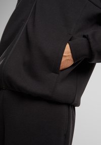Chaqueta negra de tejido suave con cremallera frontal, mangas raglán y bolsillos laterales; combinada con pantalones a juego en el mismo material.