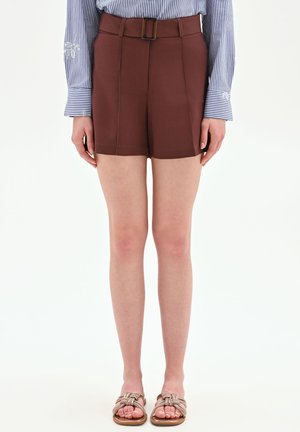 Donna che indossa pantaloncini marroni a vita alta con cintura, camicia a righe blu e bianche a maniche lunghe e sandali bassi con cinturini intrecciati.