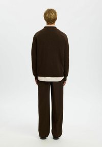 Brauner, gestrickter Cardigan mit lockerem Schnitt, flachem Kragen und geripptem Saum, über einem hellen, kragenlosen Hemd getragen und kombiniert mit dunklen Hosen.