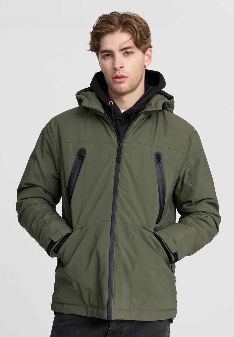 Giubbotto imbottito verde oliva con cappuccio, dotato di tasche sul petto con zip, cerniere nere a contrasto e polsini a coste. Texture liscia.