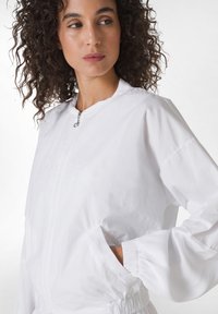 Jachetă bomber albă realizată dintr-un material ușor, având închidere cu fermoar, manșete elastice și buzunare laterale. Textură netedă, design casual.
