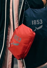 Dvě nadýchané tašky visící; jedna červená se šedým textem "1853" a vzorem, druhá tmavě modrá se podobným textem. Tašky mají zipové uzávěry a texturu látky.