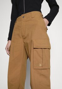 Pantalons cargo en tissu beige à coupe décontractée, avec des poches latérales et un logo brodé sur la cuisse. Texture lisse et conception robuste.