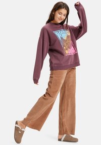 Bordeaux kleurig sweatshirt met grafisch ontwerp, gecombineerd met lichtbruine corduroy wijde broek en bruine instappers met bontaccenten.