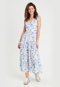 Robe blanche avec imprimé floral et paysager bleu, décolleté en V, manches volantes et jupe à volants, associée à des baskets métalliques.