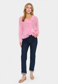 Blouse en voile rose avec détails à volants, décolleté en V, manches longues, associée à un jean bleu foncé à revers et des ballerines à motifs légers.