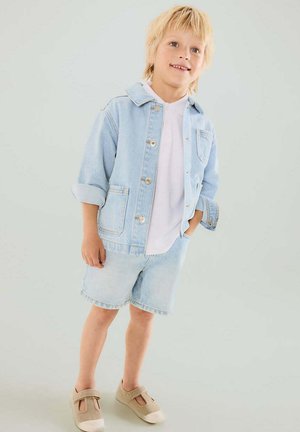 Enfant blond souriant debout avec une main dans la poche, portant une veste en jean claire, une chemise blanche, un short en jean et des chaussures beiges à brides.