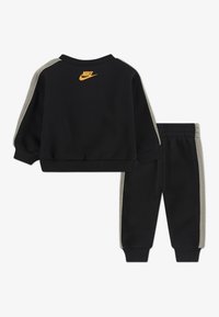 Crni Nike dukserica i hlače sivo-smešnih stranica. Sadrže rebrasta završetka na rukavima i struku. Na leđima se nalazi narančasti logo u gornjem centru.