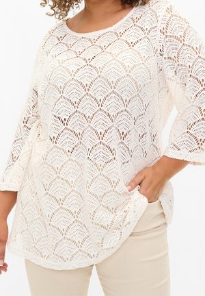 Kobieta w kremowej, wzorzystej koronkowej bluzce z rękawami do połowy przedramienia oraz beżowych spodniach, z dłońmi spoczywającymi w kieszeni.