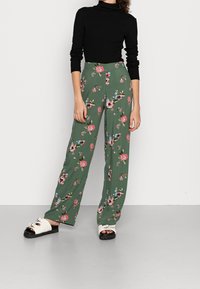 Groene wijde broek met bloemenmotief en hoge taille, met meerkleurige bloemen. Gecombineerd met een zwarte coltrui en witte instappers.