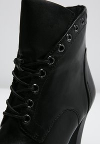 Botín de cuero negro hasta el tobillo con un tacón alto, punta redondeada, cierre de cordones y detalles de ojetes a lo largo del costado. Textura suave.