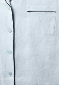 Lyseblå hørskjorte med en lomme og sorte pipings langs kanterne. Knapperne er blå, hvilket tilføjer en subtil kontrast til stoffet.