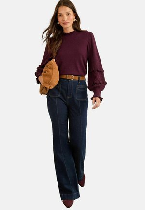 Burgundy textured long-sleeve top with ruffled sleeves, paired with dark denim wide-leg jeans and a brown belt, holding a brown handbag.