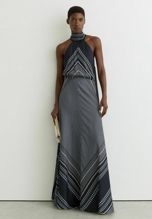 Kobieta w czarno-białej maksi sukience na szyję z wzorem, trzymająca małą beżową kopertówkę, stojąca na tle białej ściany.