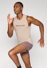 Beige ärmelloses Sporttop aus strukturiertem Stoff mit braunem Logo; kombiniert mit braunen Shorts, in einer Laufpose dargestellt.