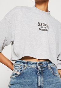 Camiseta gris cortada con cuello redondo que presenta el texto en negro "SAN DIEGO 1995 CALIFORNIA". Combinada con jeans de tiro alto azules.