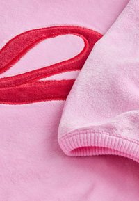 Różowa polarowa odzież o puszystej teksturze z haftowanym czerwonym wzorem. Rękawy kończą się różowym ściągaczem.