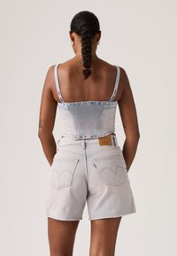 Γυναίκα με πλεξούδες, φοράει σορτς από ανοιχτό τζιν ύφασμα και ταιριαστό εφαρμοστό crop top, στέκεται με τα χέρια πίσω από την πλάτη μπροστά σε απλό φόντο.