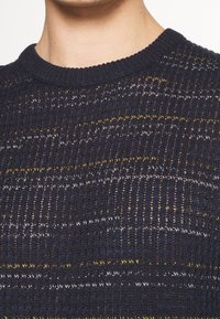 Pull en maille bleu marine à col rond ; présente des rayures texturées jaunes et blanches, créant un motif sur le tissu.