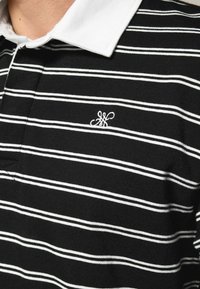 Sort-hvidstribet polo skjorte med hvid krave; har et lille hvidt logo på brystet; lavet af tekstureret stof.