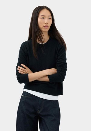 Vrouw met lang donker haar, die een zwarte trui draagt over een wit overhemd en een donkere spijkerbroek, staat met gekruiste armen en kijkt opzij.