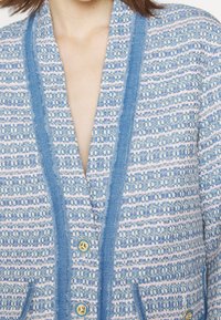 Femme portant une veste texturée bleu et blanc à motifs avec un liseré bleu et des boutons dorés, partiellement ouverte au niveau de la poitrine.